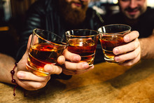 Cut View Of Three Bearded Young Men Holding Glasses With Rum Together. They Smile. People Sit In Bar.