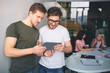 © estradaanton - Two young and strong young men stand together and look at tablet. They are interested. Young women sit at table behind them and talk. People are in one white room.