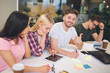 © estradaanton - Handsome young bearded man sit among his coworkers and hold big thumb up. He smiles on camera. Other people work and look down. They all are in one room.