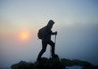 © anatoliy_gleb - Silhouette of tourist hiker man with backpack and trekking sticks on rocky mountain peak on copy space background of foggy cloudy misty blue sky and raising bright orange sun at dawn.