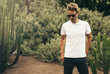 © J-Project - Young handsome bearded guy wearing a blank white t-shirt and sunglasses is standing in the garden background next to a big cactus plant. Horizontal mock up style