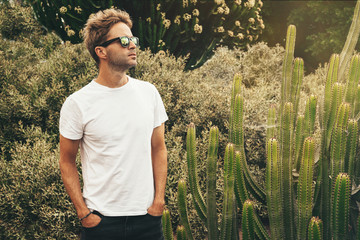 Young handsome bearded guy wearing a blank white t-shirt and sunglasses is standing in the garden background next to a big cactus plant. Horizontal mock up style