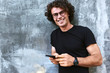 © iuricazac - Portrait of smiling man standing outdoors texting on mobile phone. Young male with curly hair wears spectacles resting outside in the city browsing on his cell phone on concrete gray background.