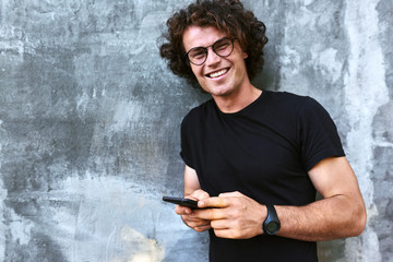 Wall Mural - Portrait of smiling man standing outdoors texting on mobile phone. Young male with curly hair wears spectacles resting outside in the city browsing on his cell phone on concrete gray background.