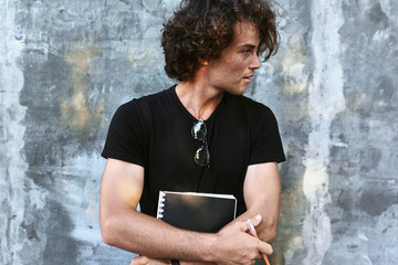 Poster - Attractive young man with curly hair, writing some notices on paper and preparing for exams, wearing black t-shirt looking at one side and posing on concrete wall.