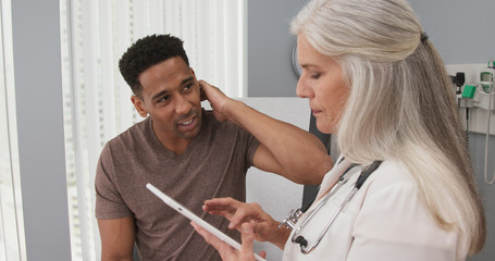 Wall Mural - Attractive black male patient paying doctor visit with neck pain. Close up of senior caucasian doctor using portable tablet to take notes of patient health