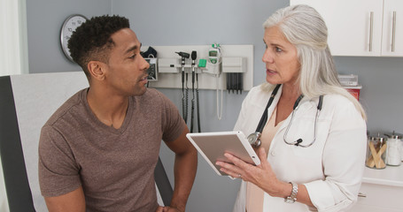 Portrait of young black man consulting with mature female doctor indoors medical clinic. Close up of senior caucasian doctor using tablet computer to share test results with male black patient