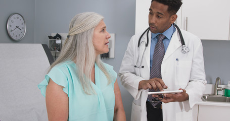 Male millennial MD taking notes on electronic tablet while listening to senior female patient. Elderly woman explaining health condition to young doctor while he uses computer pad