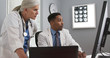 © Mark Adams - Close up of two doctors looking at computer monitor comparing notes. Young black doctor typing on laptop computer while consulting with white senior colleague