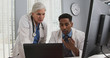 © Mark Adams - Young male black doctor talking on smartphone while working with colleague on laptop computer. Two medical doctors using technology inside office