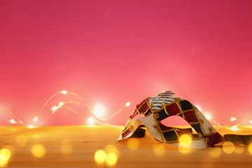 photo of elegant venetian, mardi gras mask over wooden table and pink background with garland gold lights.