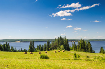 Wall Mural - beautiful summer landscape with sloping meadow, hay, forest and river with blue sky with clouds.