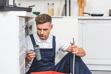 Wall Mural - thoughtful adult repairman sitting under sink and holding pipes for repairing at kitchen