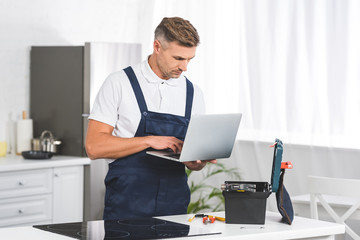 Wall Mural - thoughtful adult repairman using laptop while repairing electric stove at kitchen