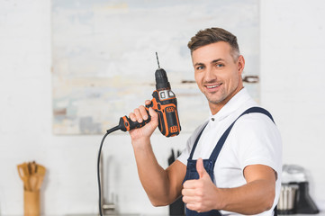 Wall Mural - selective focus of smiling adult repairman holding drill in hand and showing thumb up sign