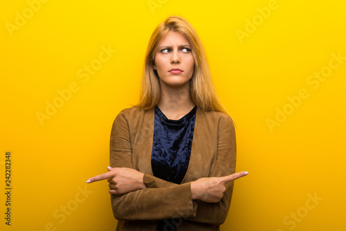 Fotografía Blonde girl on vibrant yellow background pointing to the laterals having doubts