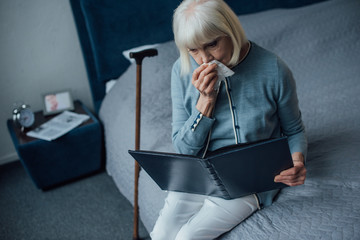 sad senior woman sitting on bed, looking at photo album and wiping tears with handkerchief at home