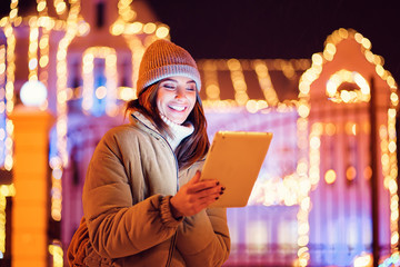 A beautiful young woman or girl in jacket and gray hat using her digital tablet in the winter. Outdoor Christmas holidays background.