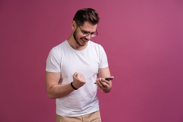 Wall Mural - Always in touch. Smiling young man holding smart phone and looking at it. Portrait of a happy man using mobile phone isolated over pink background