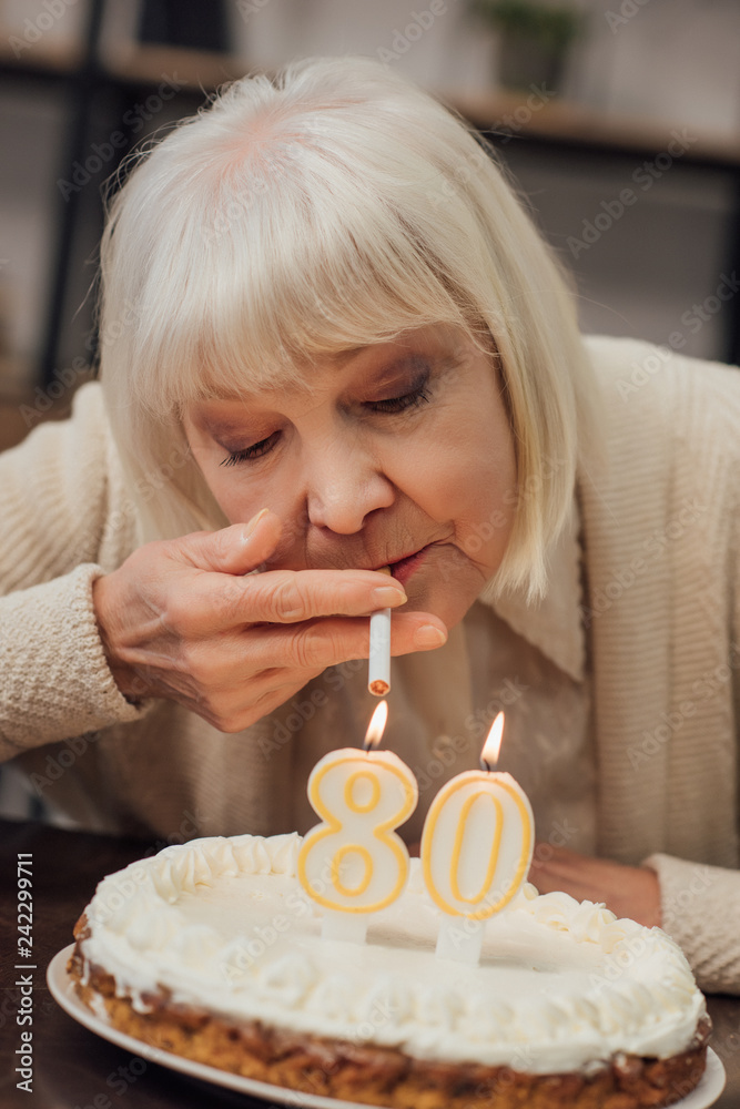 senior woman lighting up cigarette from burning candles on birthday cake at  home Stock Photo