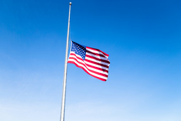 American flag at half mast against bright blue skies, signifying a day of mourning and remembrance in the United States.