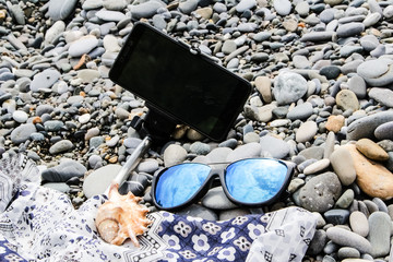 Smartphone with selfie stick, blue sun goggles and seashell lie on the seashore