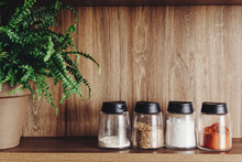 Different Spices In Glass Jars On Wooden Shelf. Luxury Modern Kitchen Furniture And Wooden Table Top. Space For Text. Spices And Green Plant On Shelf, Organization In Kitchen. Cooking Food