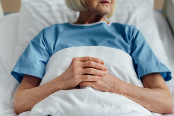 Wall Mural - cropped view of senior woman with folded hands lying in bed in hospital