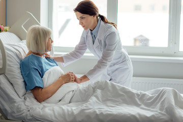 female doctor holding hands and supporting sad senior woman lying in hospital bed