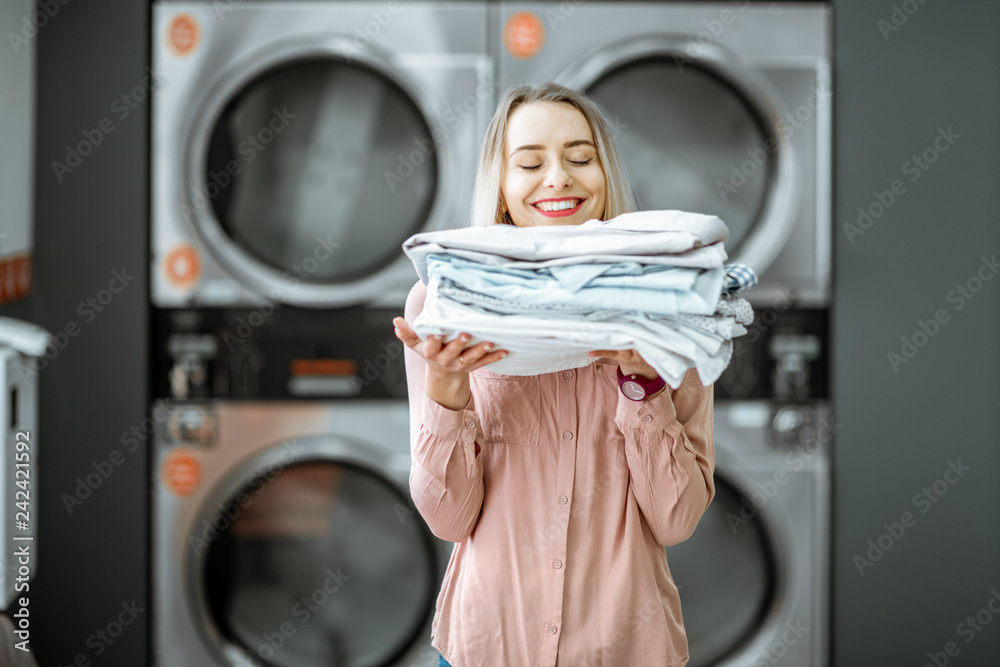 Young woman enjoying clean ironed clothes in the self serviced laundry with dryer machines on the background - obrazy, fototapety, plakaty 