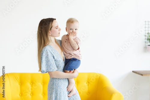 Fotografie, Obraz Adorable young blonde mother in a beautiful dress holding her little cute son in a cozy homely atmosphere with a modern interior