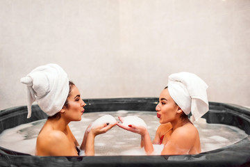 Wall Mural - Lovely young woman have some foam in hands and blow it one to another. They sit in jacuzzi with towels aroung hair. Models have rest and chill.