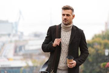 Canvas Print - Photo of european adult man 30s wearing jacket holding takeaway coffee, while walking through city street