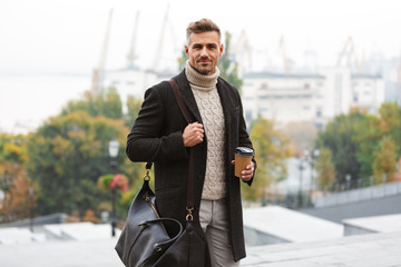 Photo of caucasian happy man 30s wearing jacket holding takeaway coffee, while walking through city street