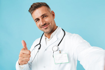 Poster - Portrait of happy medical doctor with stethoscope taking selfie photo, while standing isolated over blue background