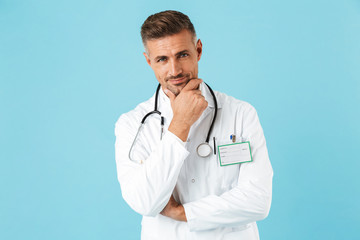 Poster - Portrait of cheerful medical doctor with stethoscope wearing white uniform, standing isolated over blue background