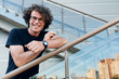 © Z A C - Smart young man smiling and wearing black t-shirt and wristwatch on the city street. Happy business male with curly hair posing for advertisement with copy space outdoor against building. People