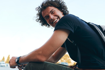 Wall Mural - Rear view of smiling cheerful young man wears black t-shirt and wristwatch on the city street. Happy male with curly hair posing for advertisement with copy space outdoor. People concept