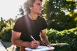 © iuricazac - Young handsome man with curly hair writting some notices on paper document and preparing for exam, sitting on the city street. Freelancer businessman makes plans for new projects.
