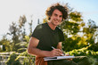 © iuricazac - Attractive man with curly hair smiling and writting some notices on paper document and preparing for exam, sitting on the city street. Freelancer businessman looking at the camera makes plans for day