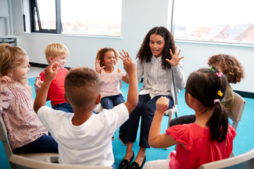 Wall Mural - A class of infant school children sitting on chairs in a circle in the classroom, raising hands and learning to count with their female teacher, close up