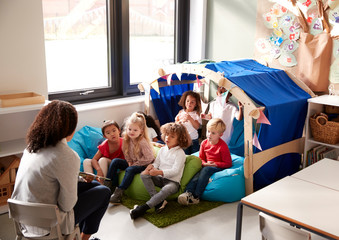 Wall Mural - Female infant school teacher sitting on a chair showing a book to a group of children sitting on bean bags in a comfortable corner of the classroom, elevated view