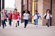 A group of smiling multi-ethnic school kids running in a walkway outside their infant school building after a lesson