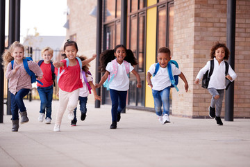 a group of smiling multi-ethnic school kids running in a walkway outside their infant school buildin