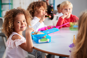 close up of smiling young children sitting at a table eating their packed lunches together at infant