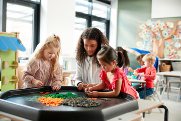 Two young schoolgirls standing at a table playing a game with their female teacher in an infant school classroom, selective focus