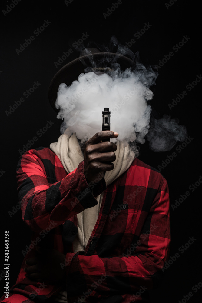 Young african man with cloud of smoke on face vape electronic cigarette  cloud by using a mod, smoke tricks of vaping device on black background.  Stock Photo