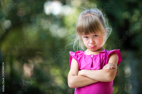 Fotografie, Obraz Portrait of pretty funny moody unhappy young blond child girl in pink sleeveless dress feeling angry and unsatisfied on blurred summer green tree copy space bokeh background