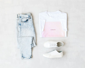 Wall Mural - White t-shirt with pink print, light blue jeans and white sneakers on grey background. Overhead view of woman's casual outfits. Flat lay, top view. Trendy hipster look.