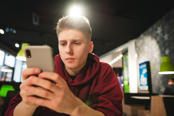 Young man uses a smartphone in a fast food cafe, is focused on looking at the smartphone screen against the backdrop of a cafe. Teen is writing a message on the smartphone.